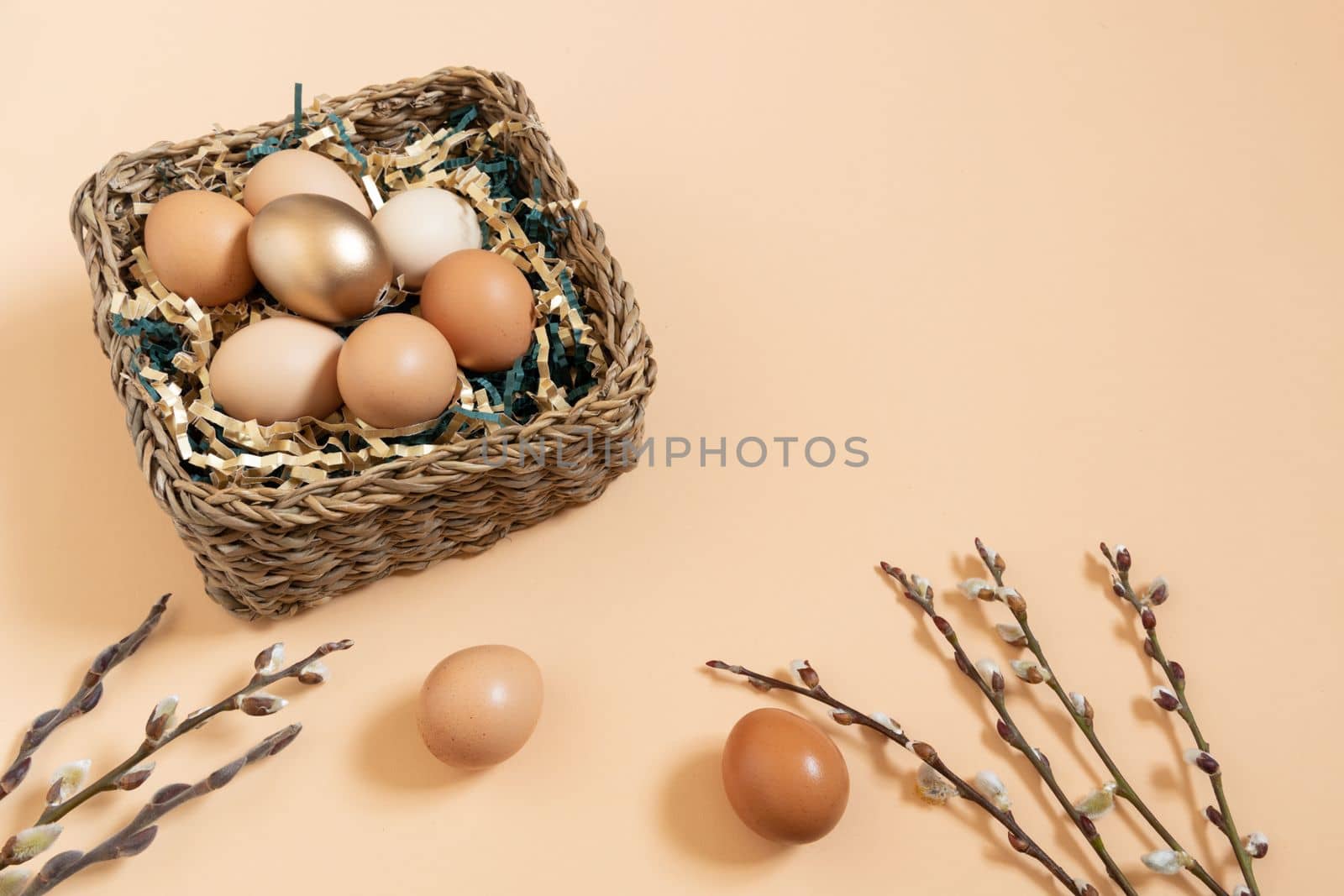 Easter eggs natural and gold in basket with paper filler . Branch of willow catkins. Light pastel orange background with copy space.