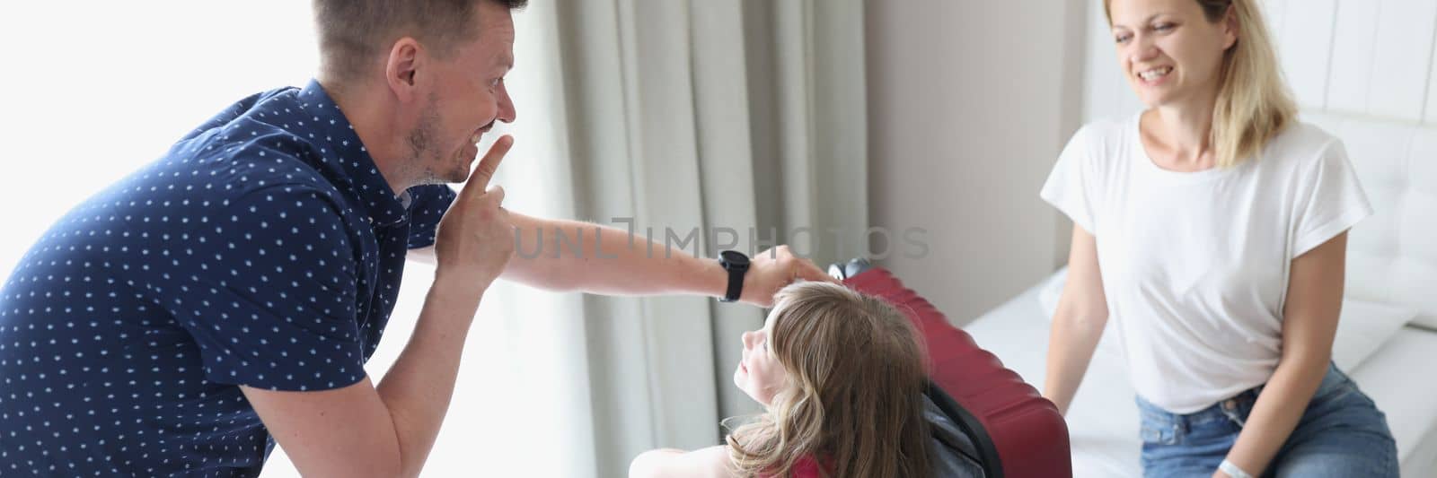 Cheerful dad and mom play hide and seek with child and suitcase. Dad asks child to sit quietly in suitcase