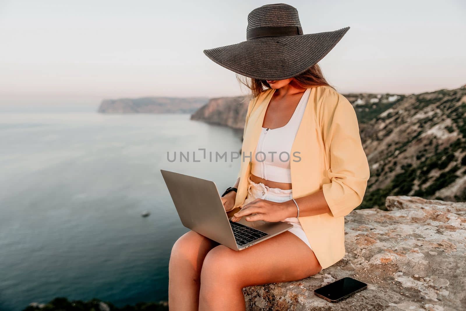 Successful business woman in yellow hat working on laptop by the sea. Pretty lady typing on computer at summer day outdoors. Freelance, travel and holidays concept.