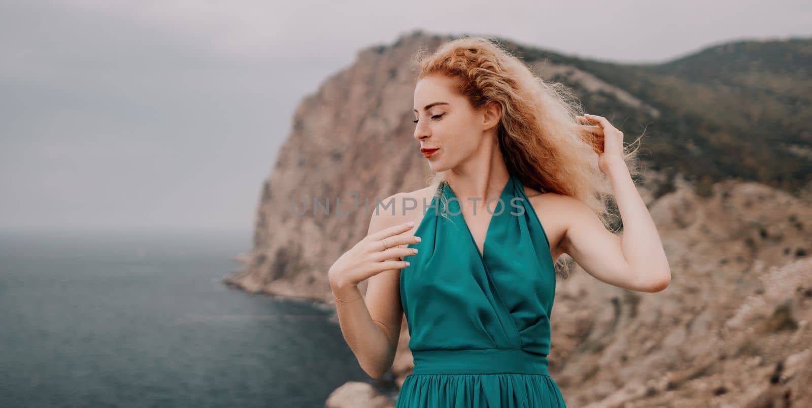 Redhead woman portrait. Curly redhead young caucasian woman with freckles looking at camera and smiling. Close up portrait cute woman in a mint long dress posing on a volcanic rock high above the sea by panophotograph