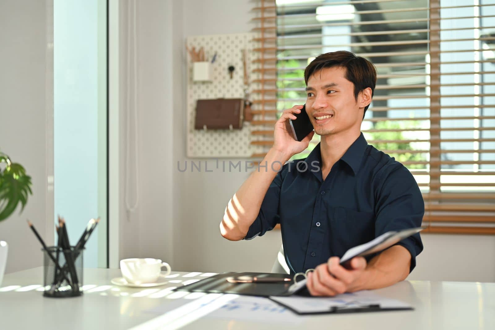 Handsome asian businessman having pleasant conversation on mobile phone with client at his office.