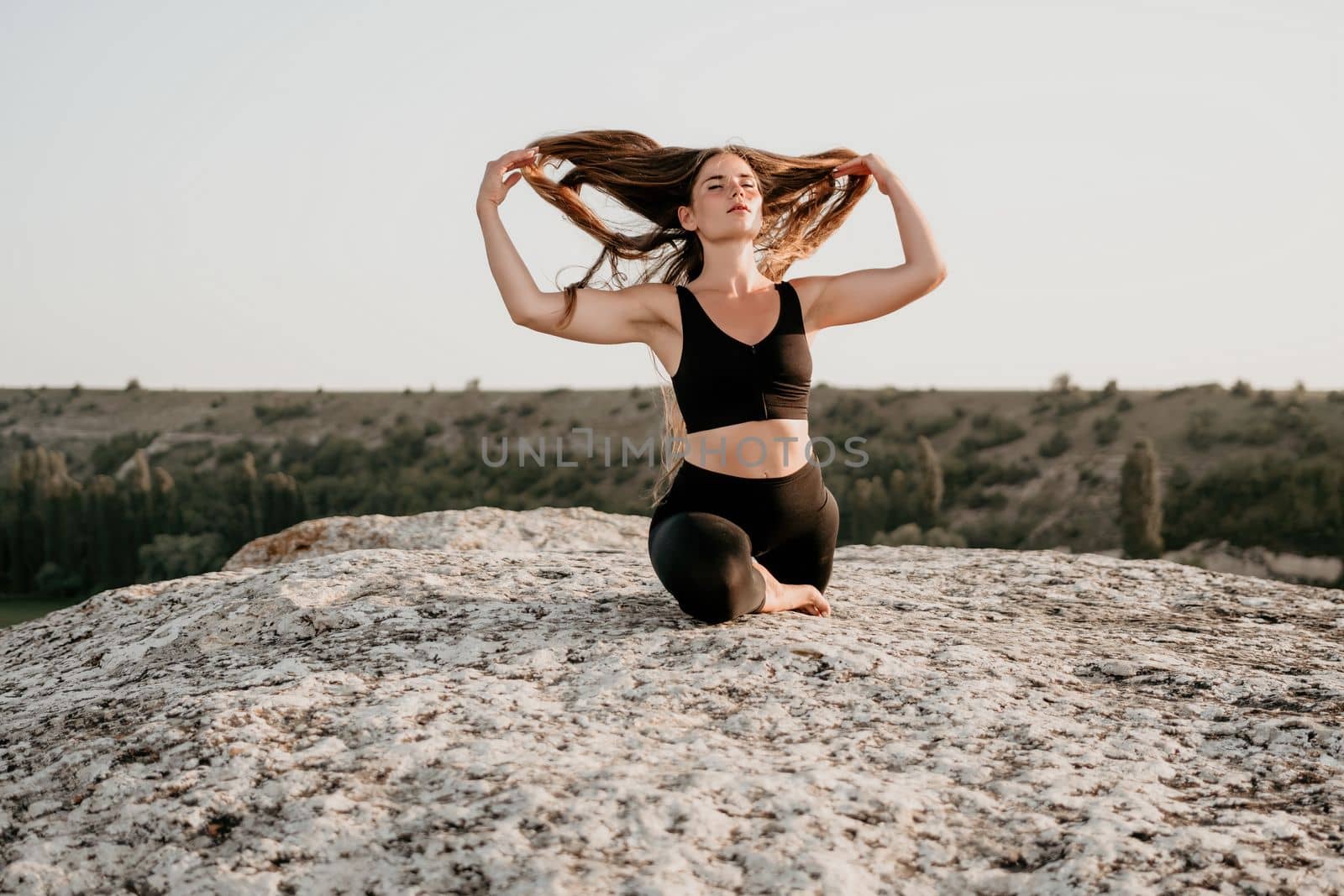 Well looking middle aged woman with long hair, fitness instructor in leggings and tops doing stretching and pilates on the rock near forest. Female fitness yoga routine concept. Healthy lifestyle.