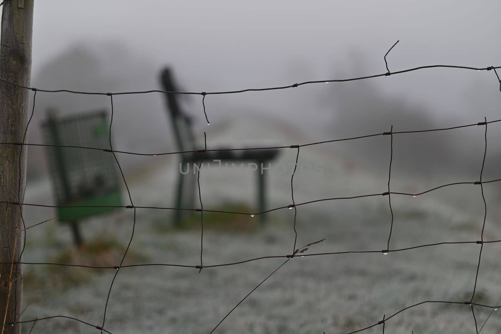 Park bench and a dustbin behind a sheep fence on a dike on a foggy morning in Wintertime