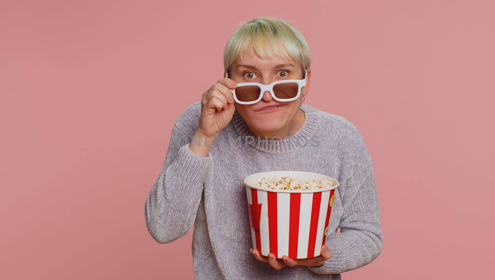 Excited woman in 3D glasses eating popcorn, watching interesting tv serial, sport game, film, online social media movie content. Young girl enjoying domestic entertainment on pink studio background