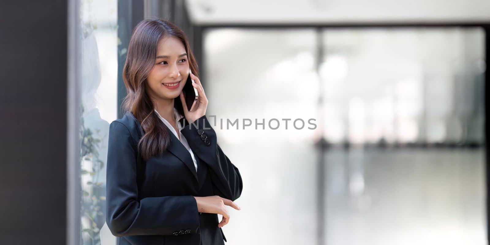 Happy young asian business woman wearing suit holding mobile phone standing in her workstation office.