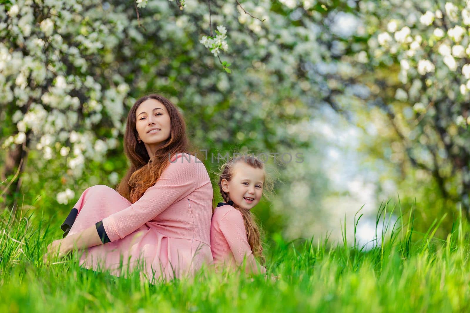 mother and daughter sit in a flowering garden with their backs to each other