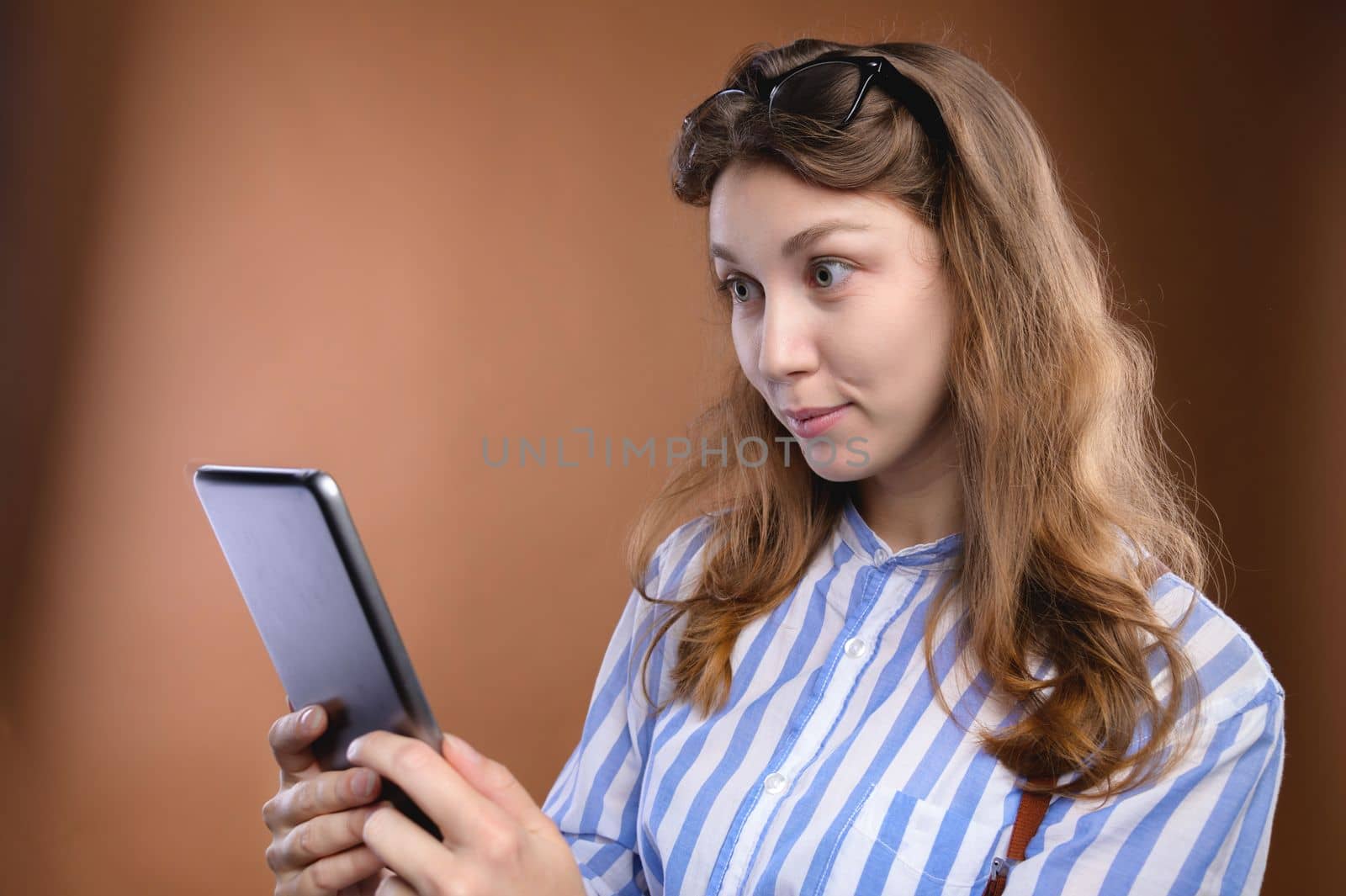 Portrait of a cheerful charming girl holding a digital tablet with a smile and looking into it. Student with glasses rejoices, studio shot.