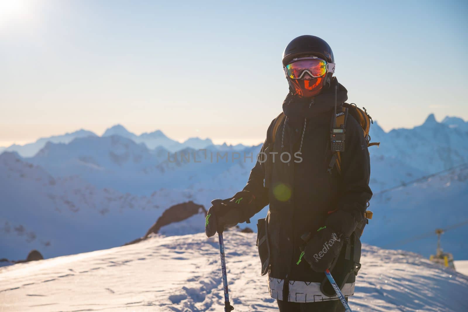 A skier in a fashionable jacket and with a backpack stands on skis against the backdrop of the Caucasus Mountains and a ski resort on a sunny day. Woman on vacation doing winter sports.