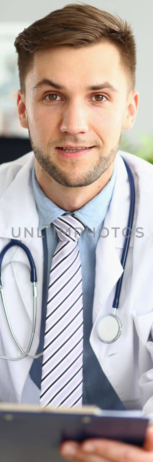 Portrait of a smiling handsome doctor working in clinic. Doctor cardiologist therapist choosing right method of treatment and medical diagnosis sitting at table with patient card
