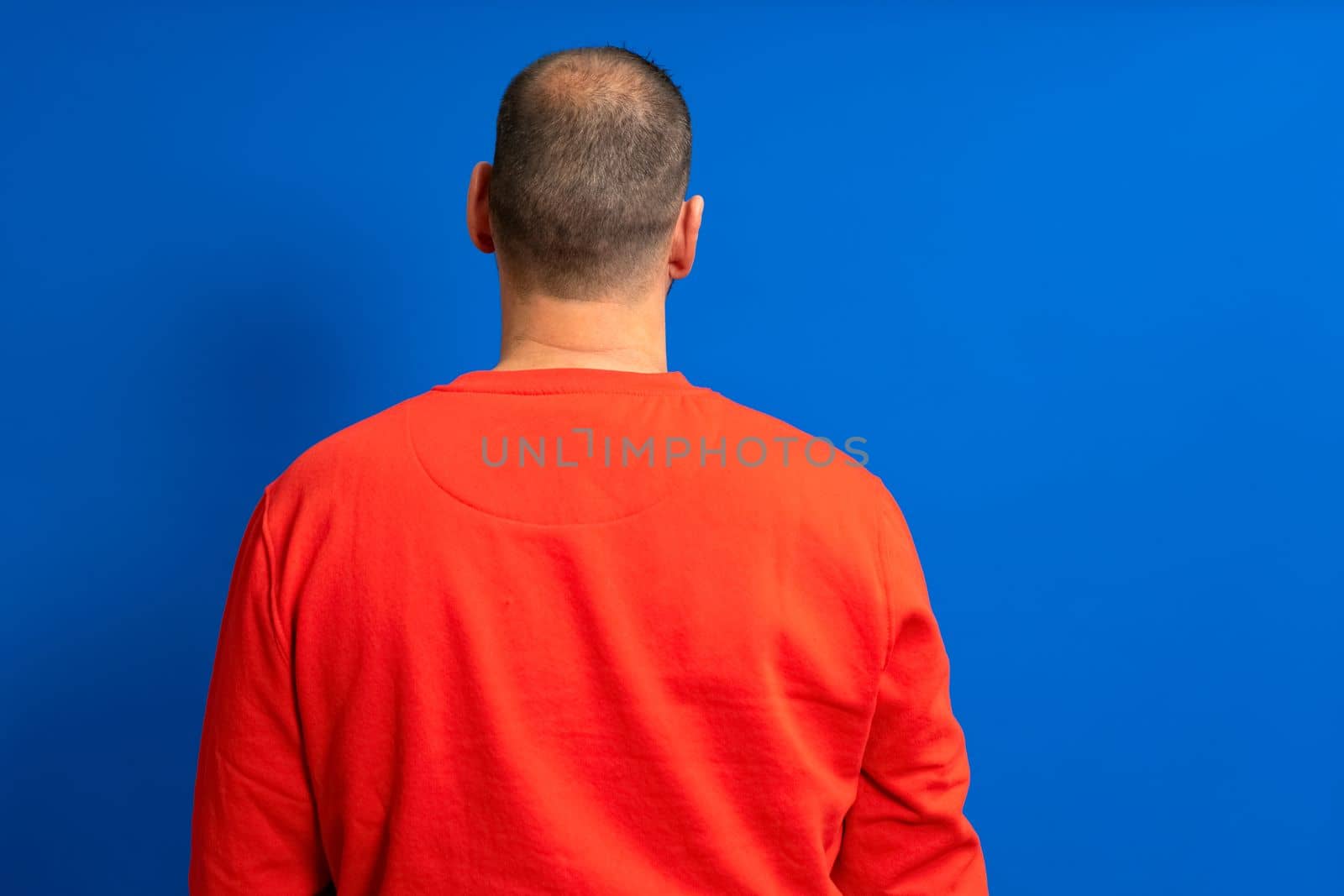 Man in his 40s with a red sweatshirt and very short hair posing with his back isolated over blue background