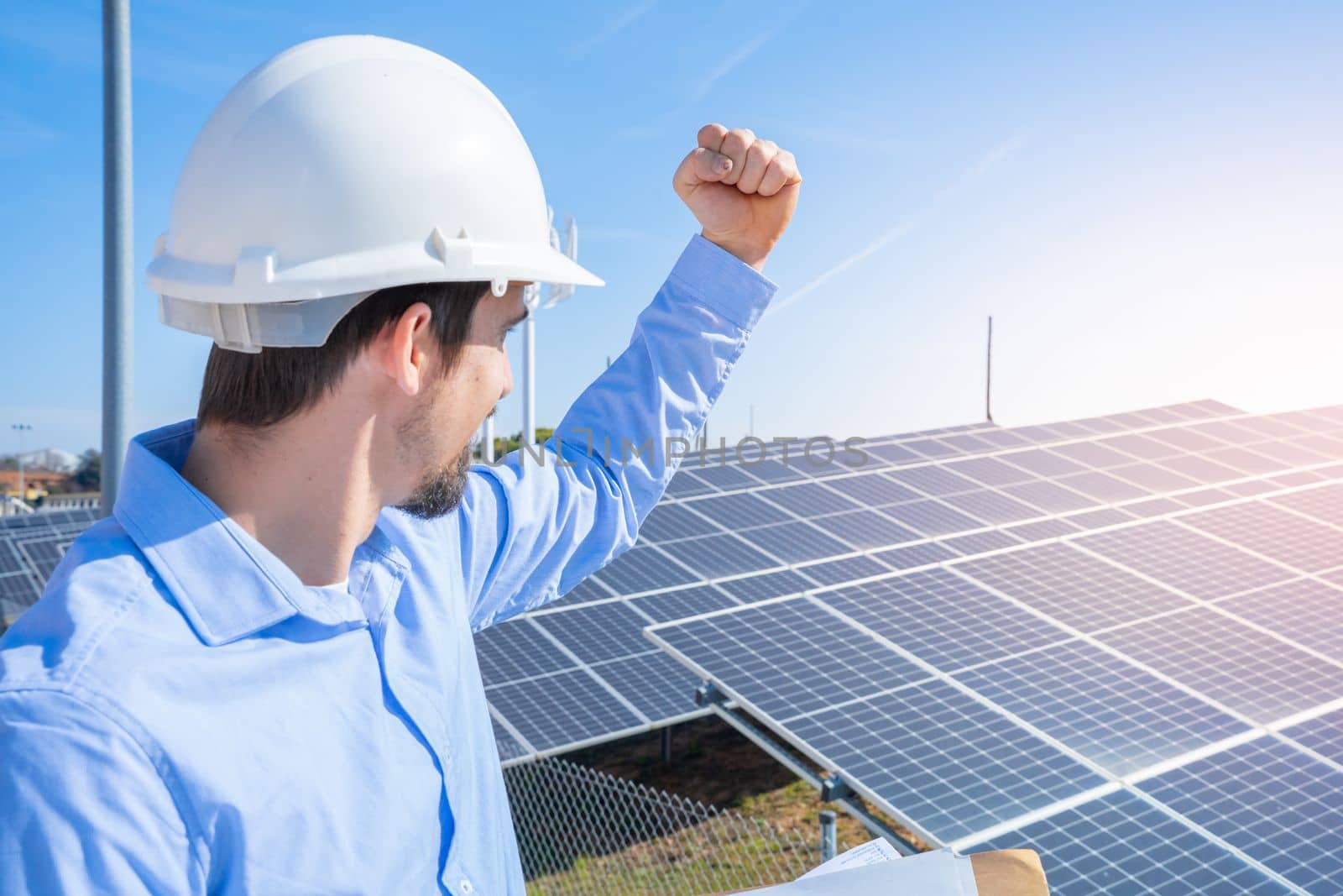 Happy engineer in protective helmet on a solar power plant celebrating success. Sun panels station development concept.