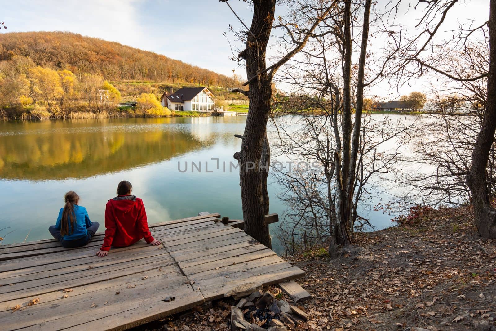 A girl and a girl are sitting on a wooden bridge by the lake and looking at a beautiful autumn landscape by Madhourse