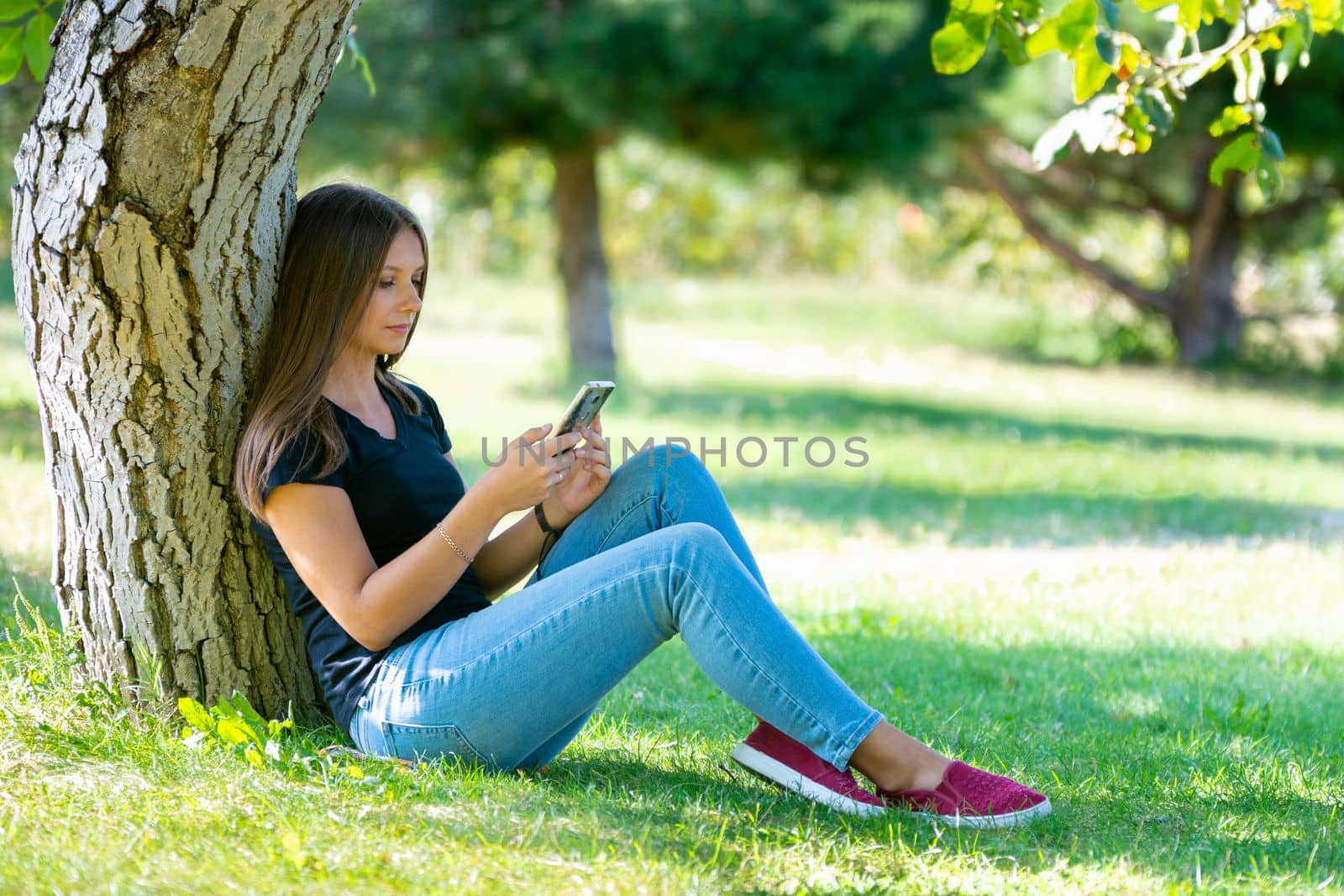A girl sits under a tree in a sunny park and looks at the smartphone screen by Madhourse