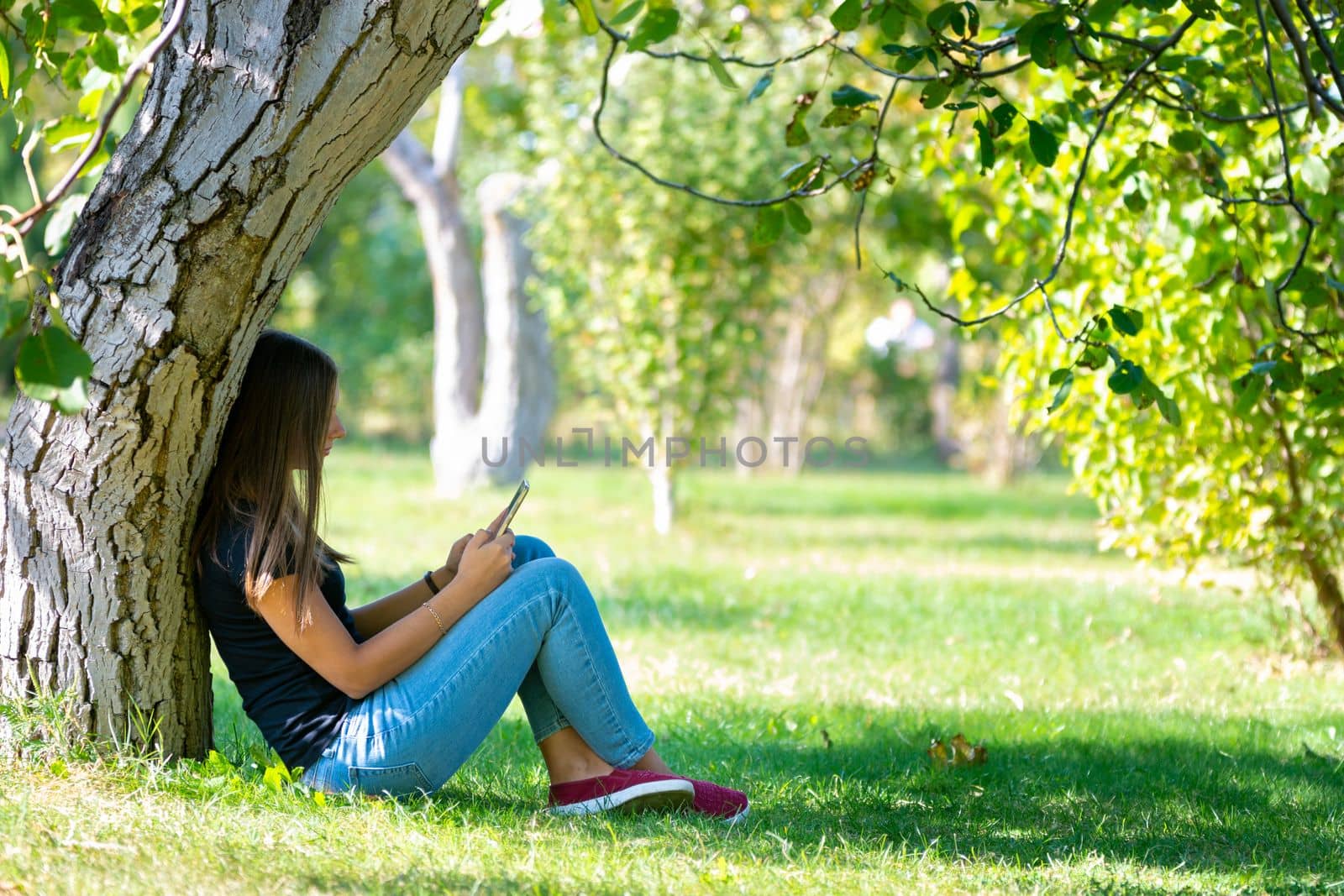 A girl sits under a tree in a sunny park and looks at the phone screen by Madhourse