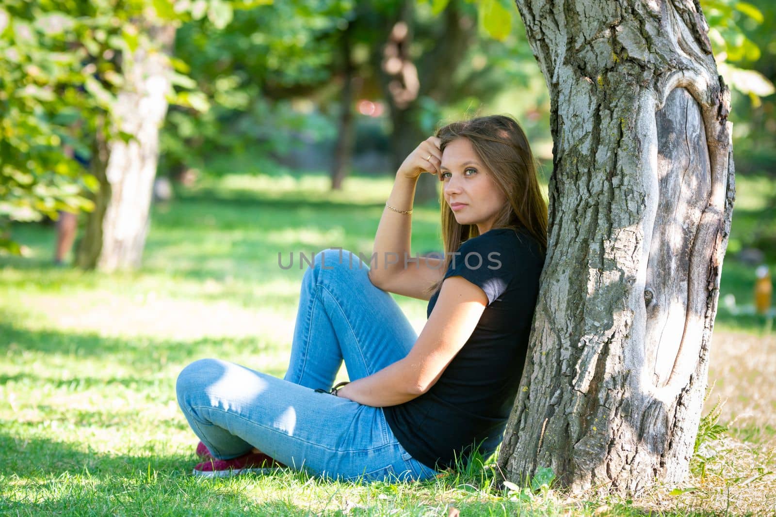 A girl sits under a tree in a sunny park and looks into the distance in thought by Madhourse