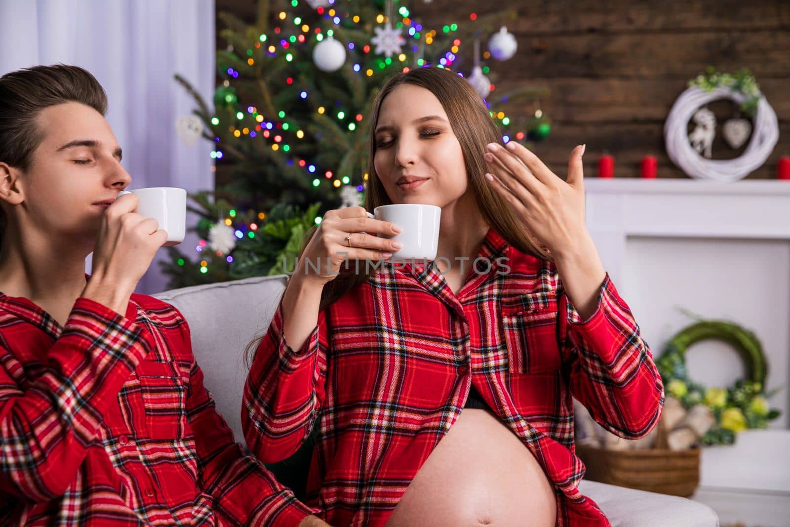 The couple sits close together with their eyes closed. Both are holding white cups inhaling the aroma of the hot drink. In the background is a fuzzy Christmas tree dressed with colorful ornaments and Christmas tree lights.