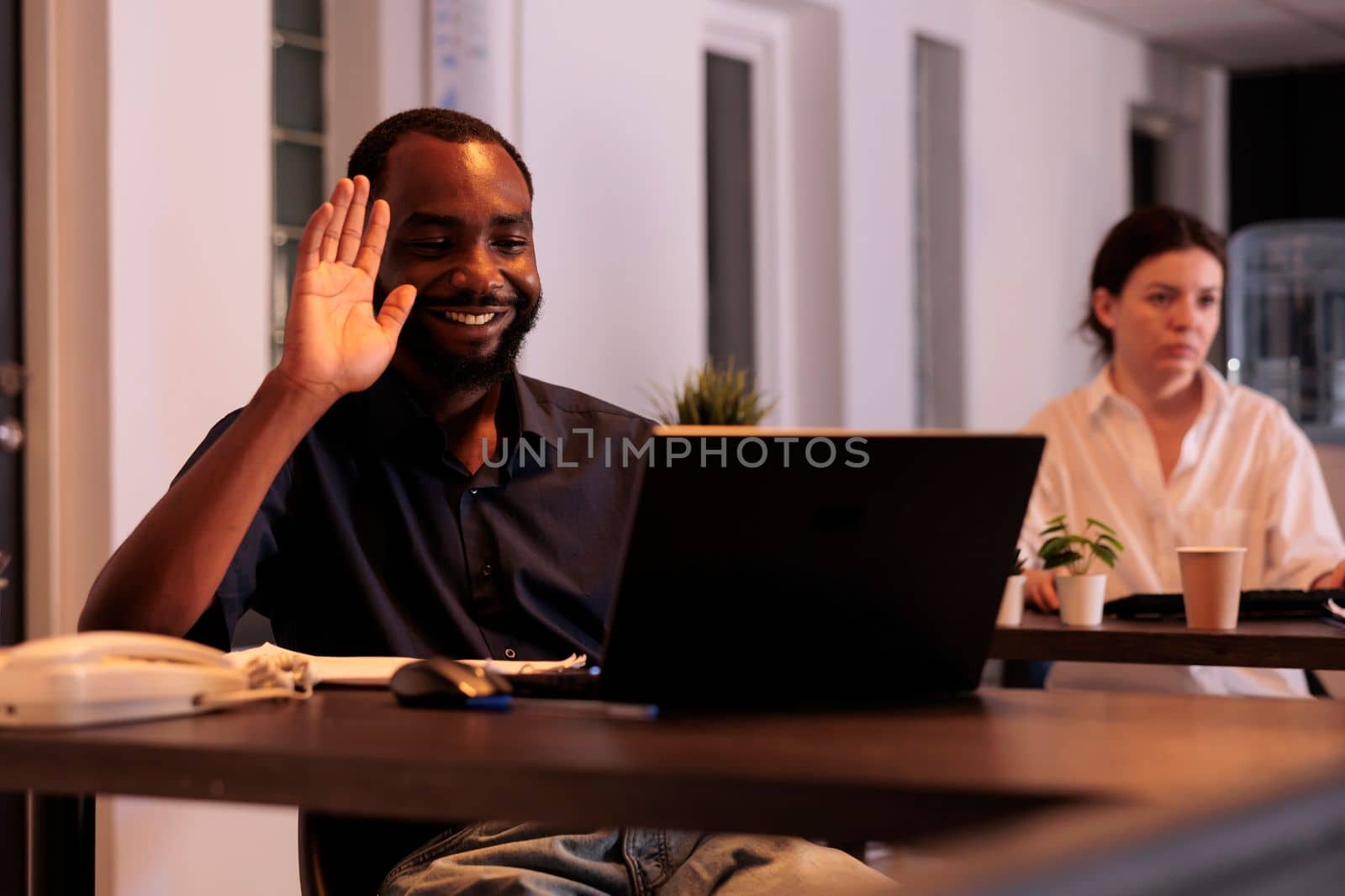 Smiling corporate worker talking on videocall, waving hi, employee answering teleconference call in office at night. African american man online communication in coworking space