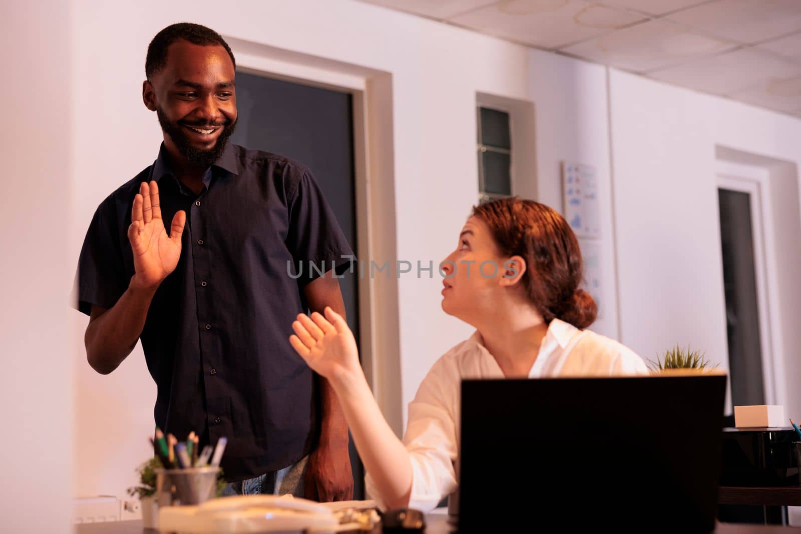 Employee meeting new colleague in corporate office, reading company report on laptop. Smiling african american man greeting employee, waving hi, standing in coworking space