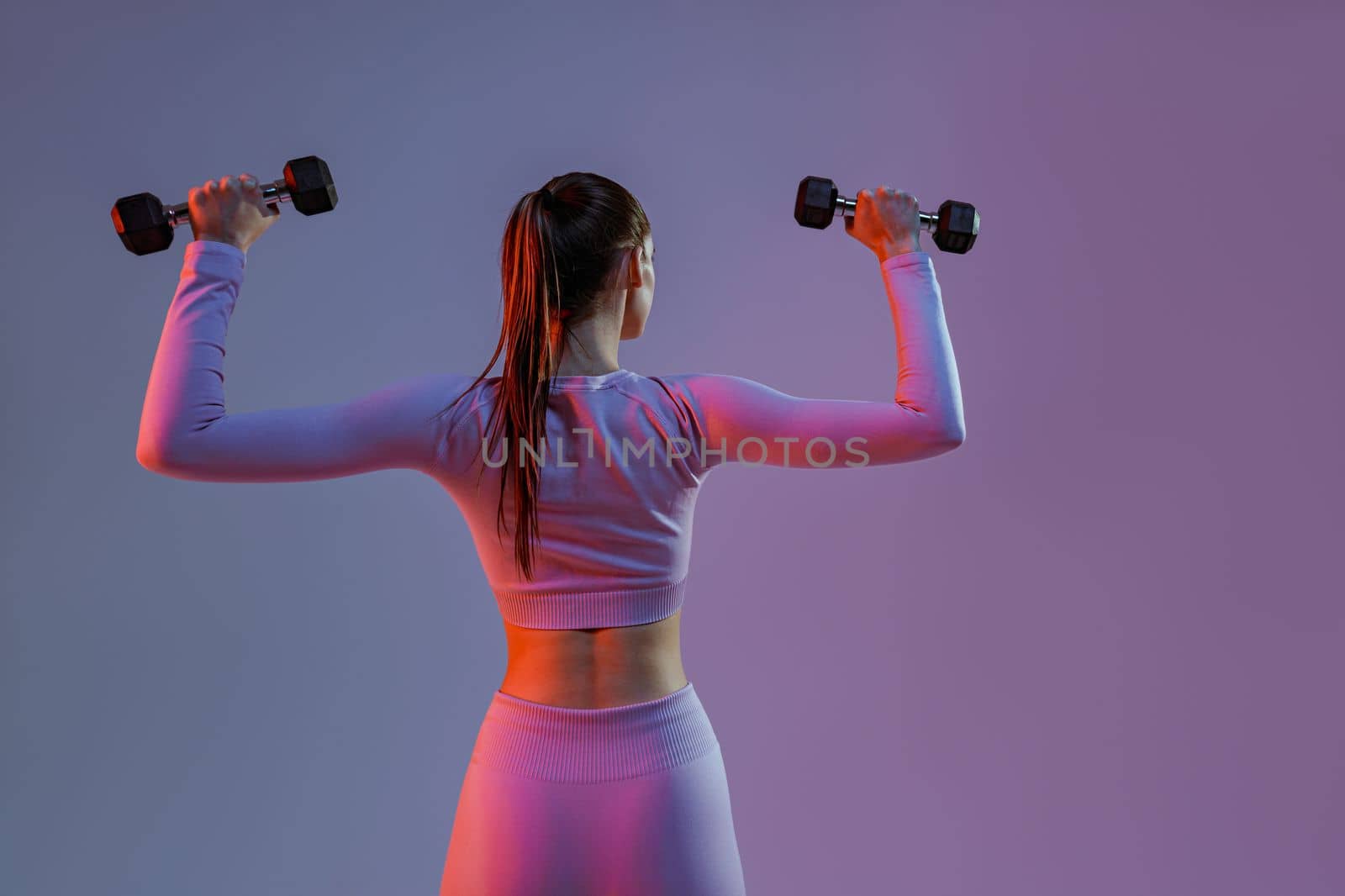 Back view of fitness woman doing exercises with dumbbells on studio background with color filter by Yaroslav_astakhov