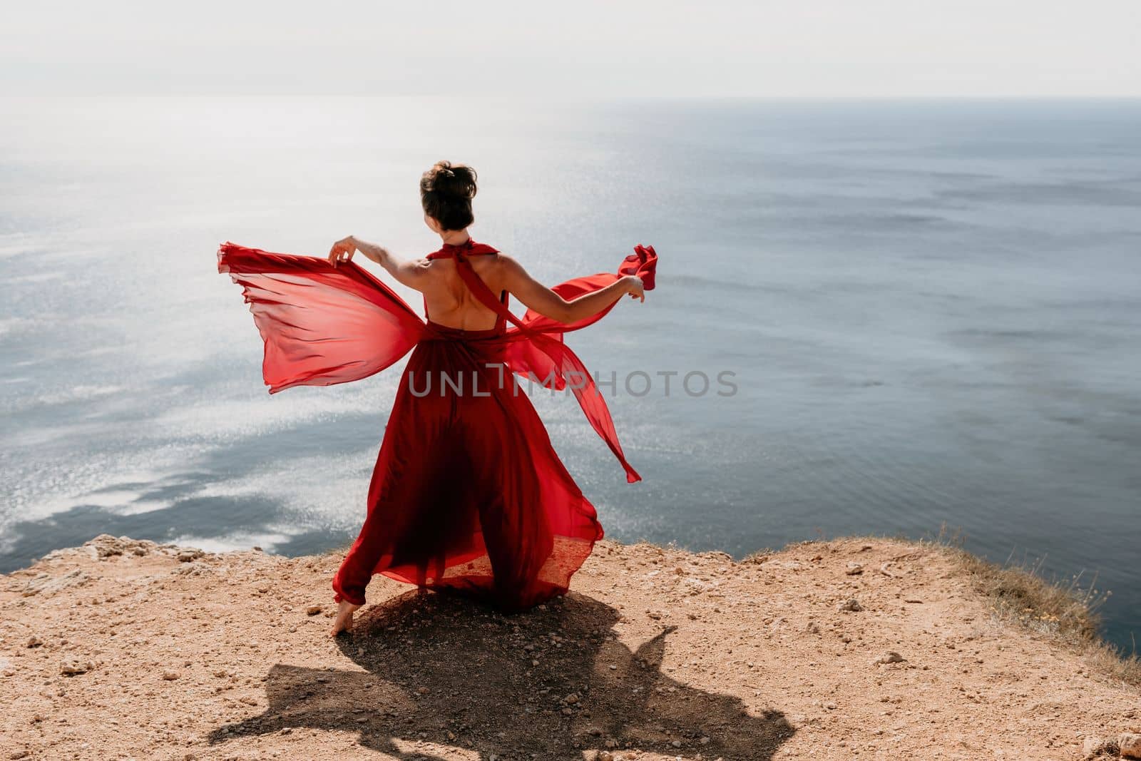 Side view a Young beautiful sensual woman in a red long dress posing on a rock high above the sea during sunrise. Girl on the nature on blue sky background. Fashion photo.