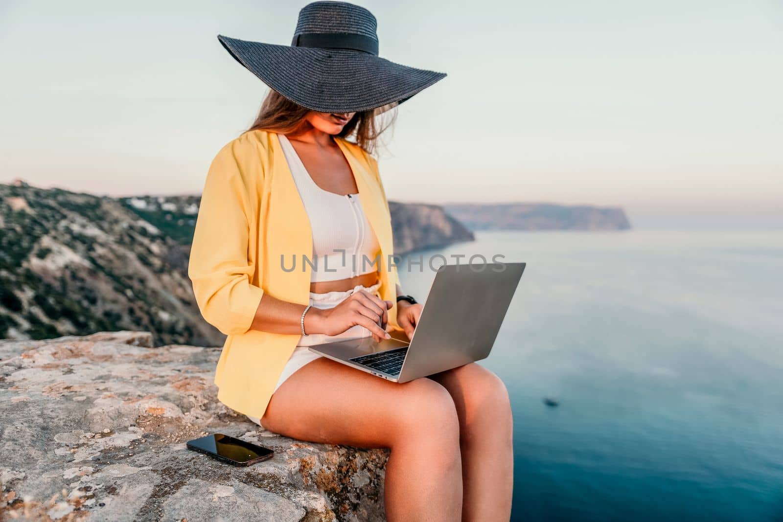 Successful business woman in yellow hat working on laptop by the sea. Pretty lady typing on computer at summer day outdoors. Freelance, travel and holidays concept.