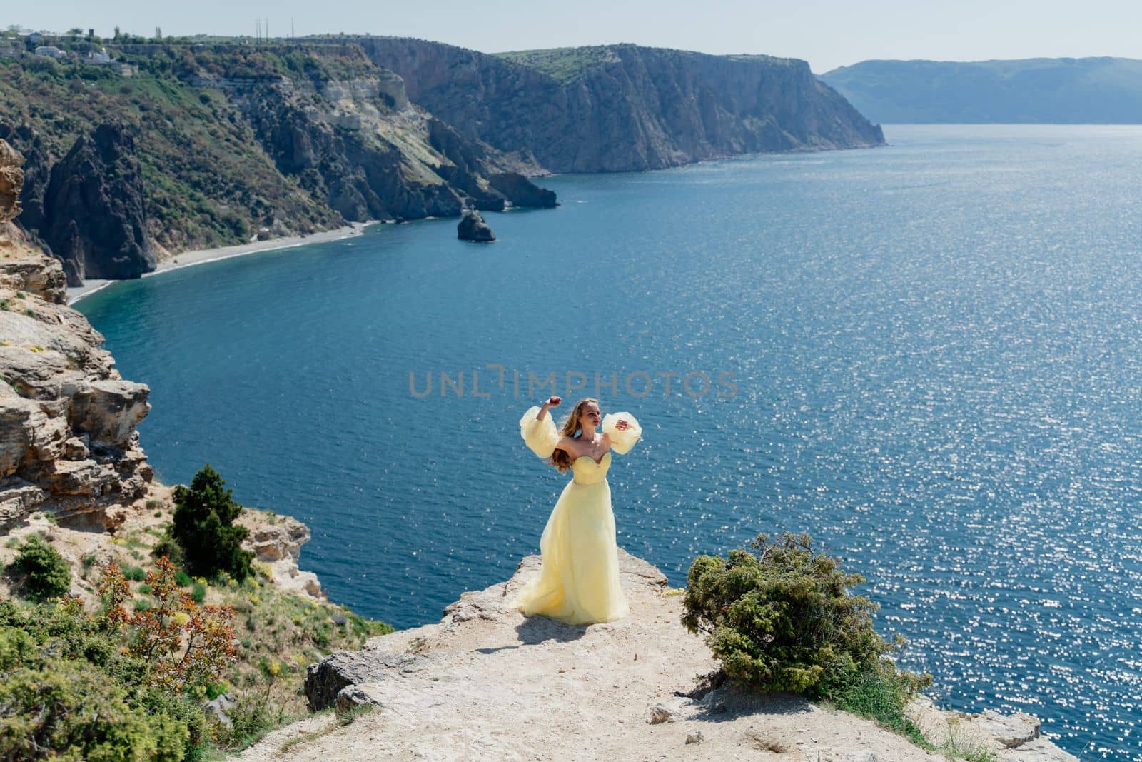 Woman in a yellow dress on the sea. Side view Young beautiful sensual woman in yellow long dress posing on a rock high above the sea at sunset. Girl in nature against the blue sky.