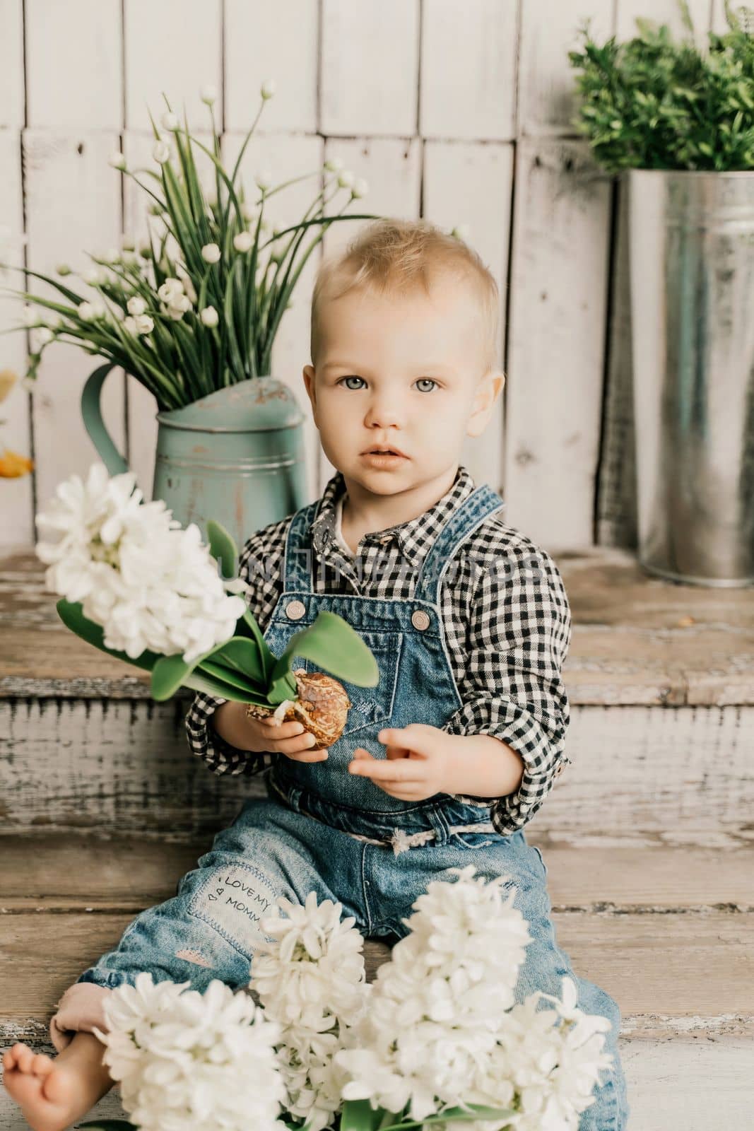 Girl 1 year old. sits holding a white hyacinth with a bulb in her hands, she is dressed in a plaid shirt and denim overalls on a wooden background with green plants in pots