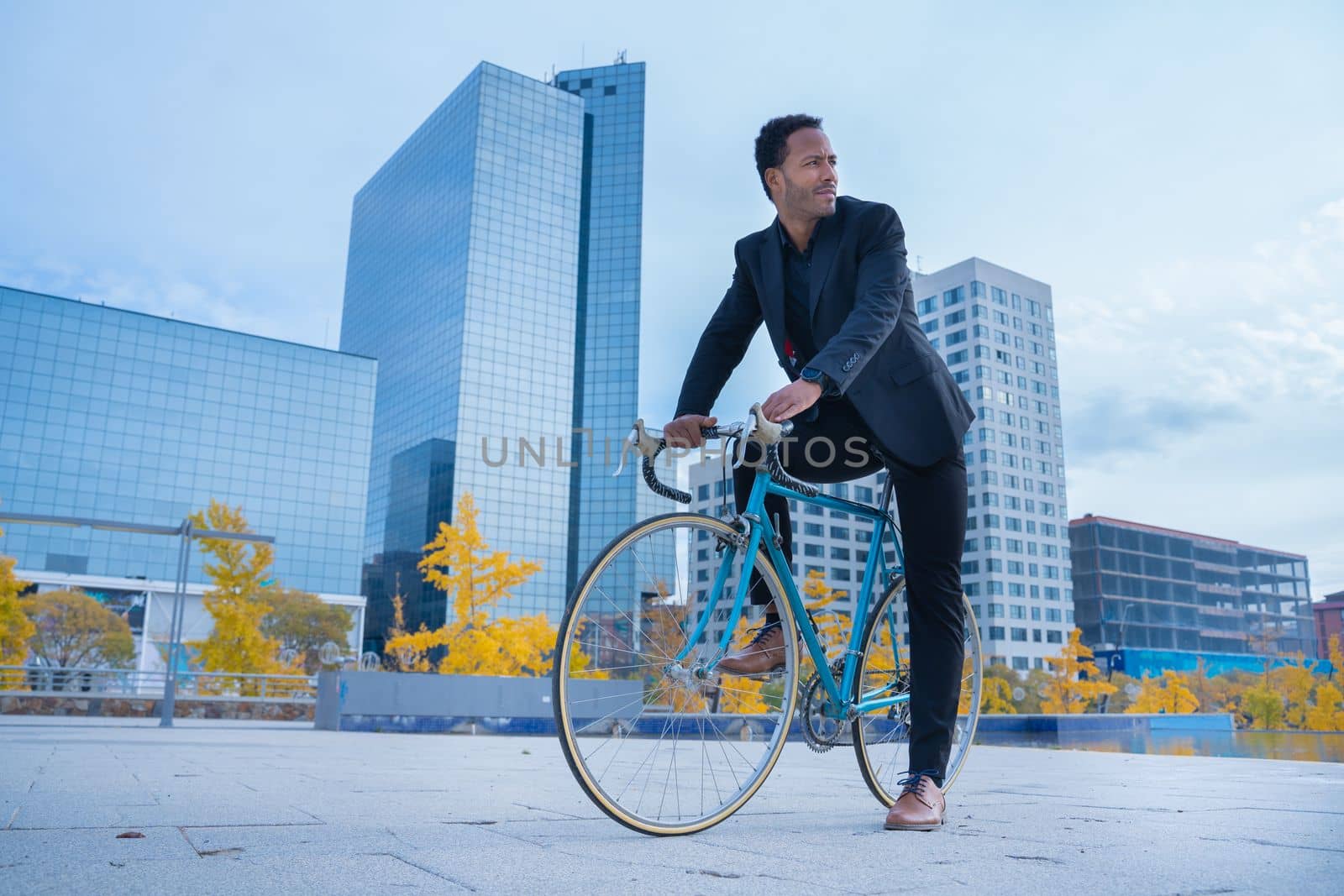 Stylish African American businessman in suit going to the office by bicycle in financial district. by PaulCarr