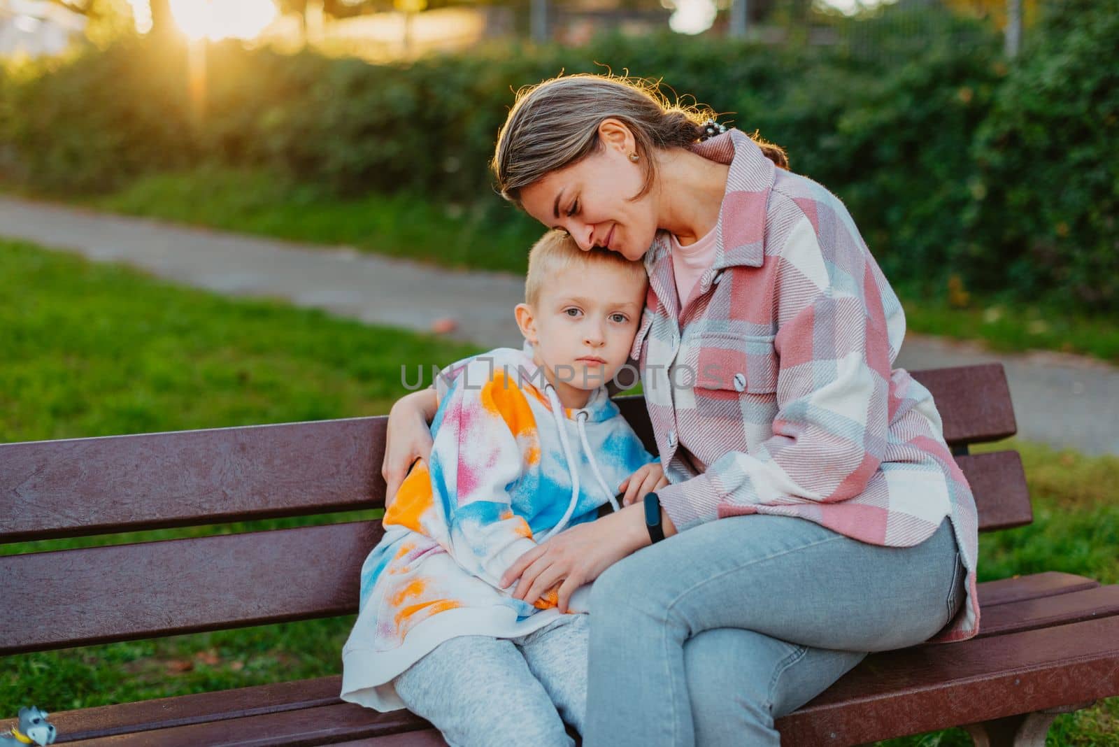 mother and son sit on a park bench in the rays of the setting sun. the concept of a family. Mother's Day. beautiful girl (mother) with a boy (son) in the park in the park are sitting on a bench at sunset.