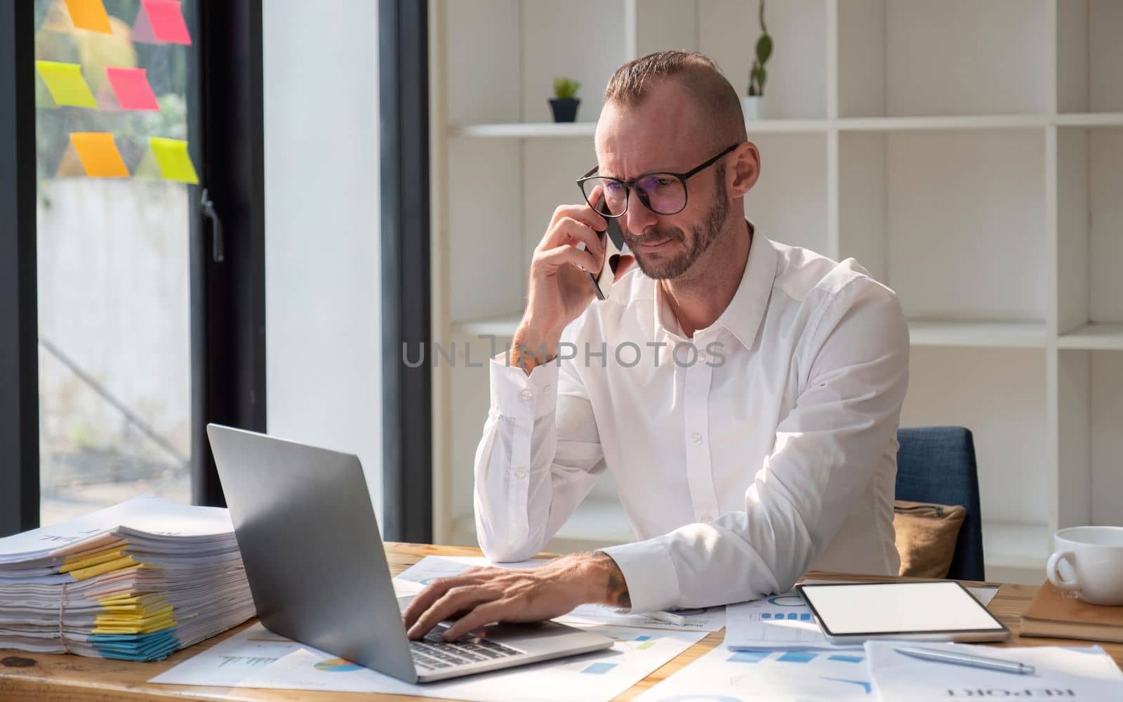 Businessman wearing glasses talking on phone, sitting at desk with laptop at office.