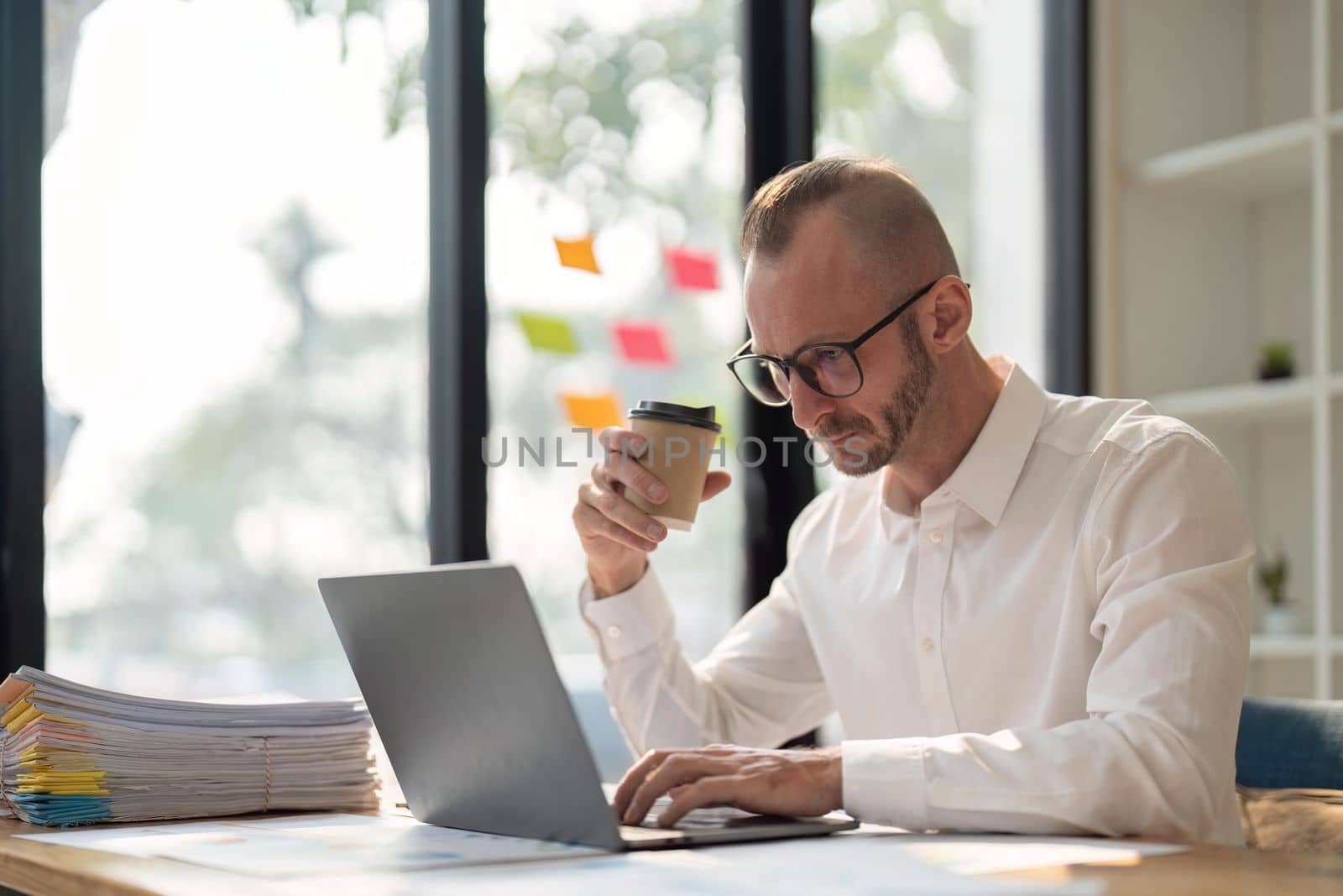 Morning coffee. young and successful business man wear eyeglasses holding cup of coffee and working with laptop while sitting at his working place by nateemee