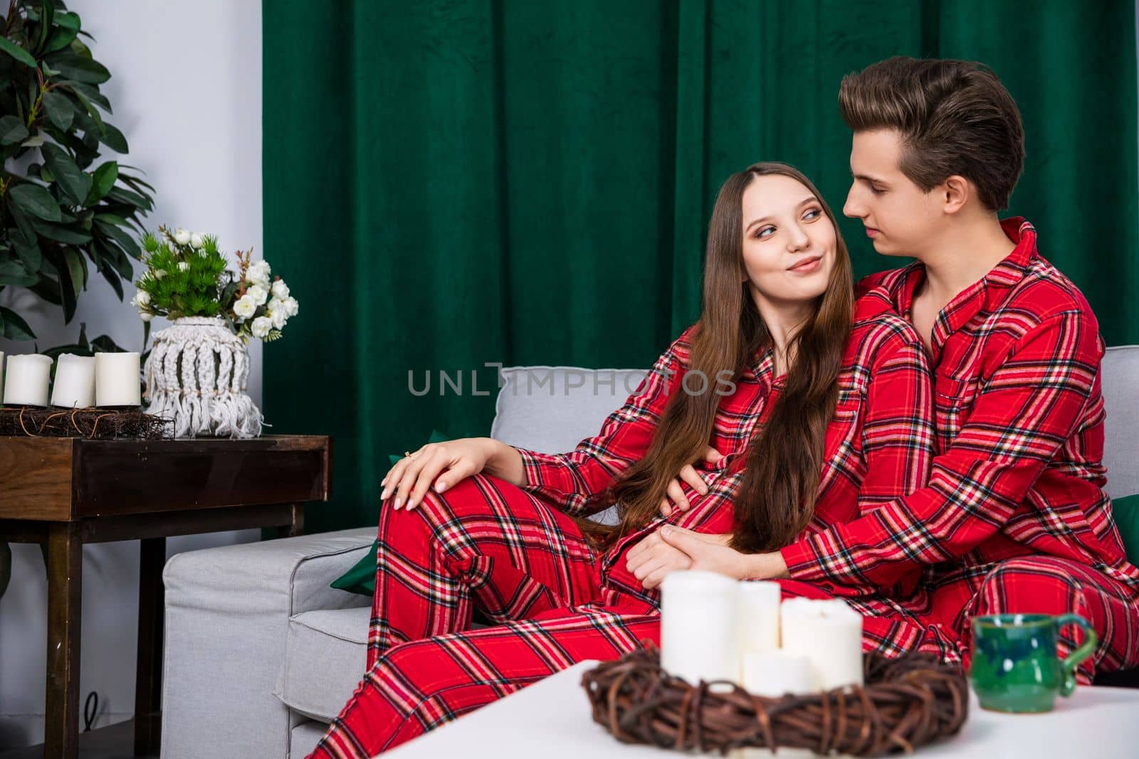 The couple in the photo shoot pose in identical red checkered pajamas. A heavy green curtain is seen in the background. The setting is romantic and in trendy colors of gray and green.