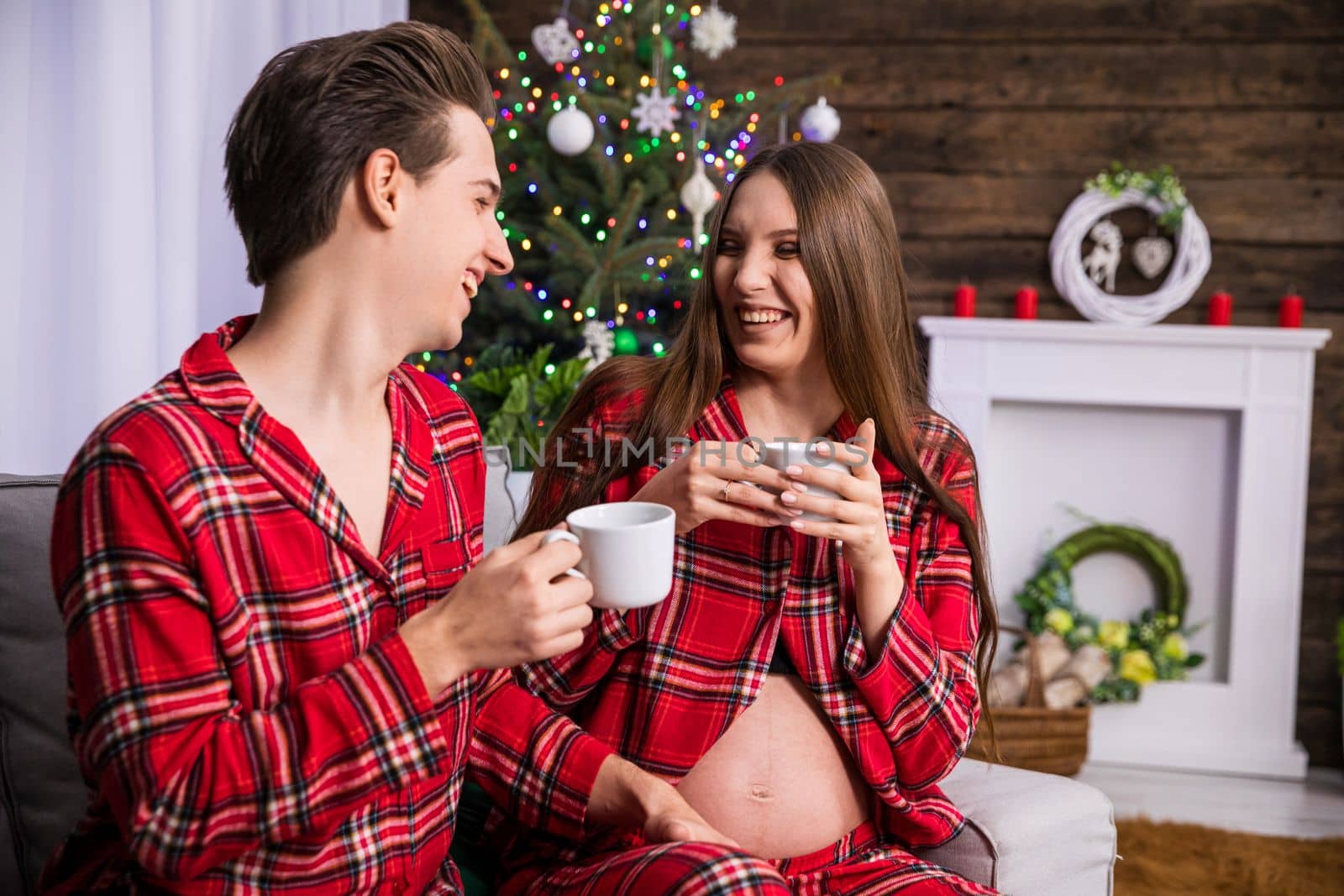 A man and a woman with advanced pregnancy sit on a couch against the background of a Christmas tree. The couple is holding white teacups in their hands. Both are facing each other, they are cheerful and laughing.