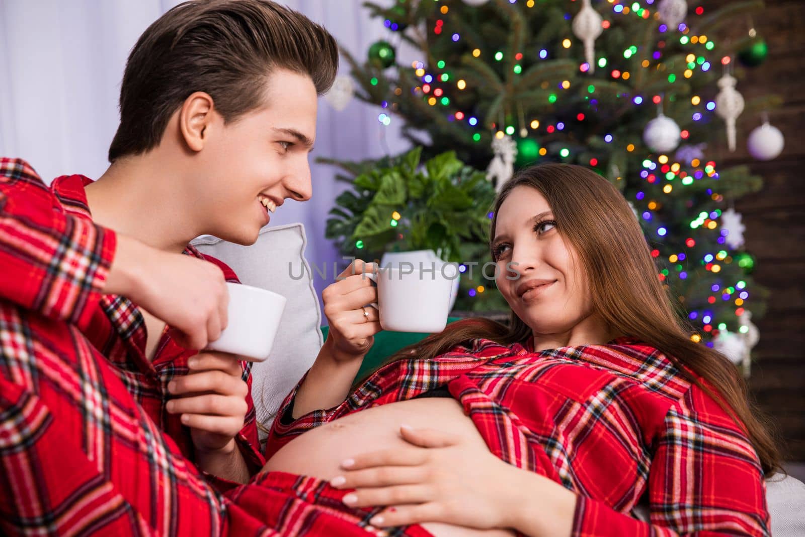 A woman in advanced pregnancy shows off her exposed pregnant belly while resting her legs on her partner's knees. The couple looks into each other's eyes and holds white cups in their hands.