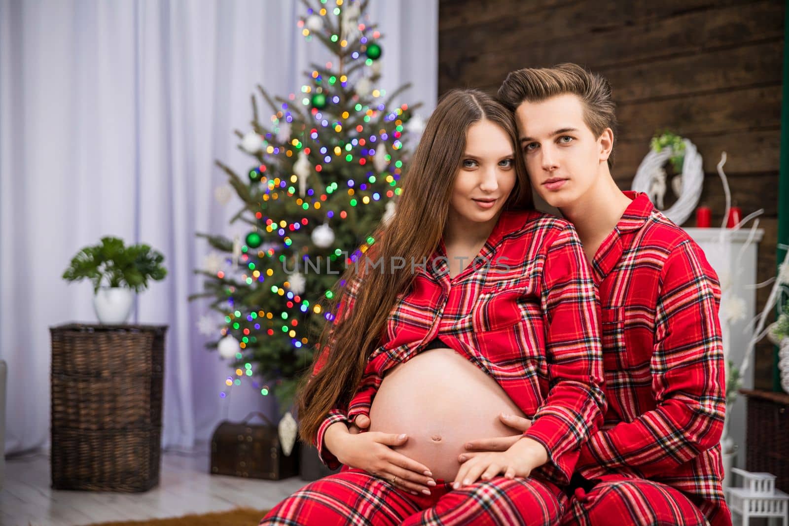 A pregnant woman and her husband sit together wearing red pajamas. A Christmas tree with lights can be seen in the background. Two people are smiling.