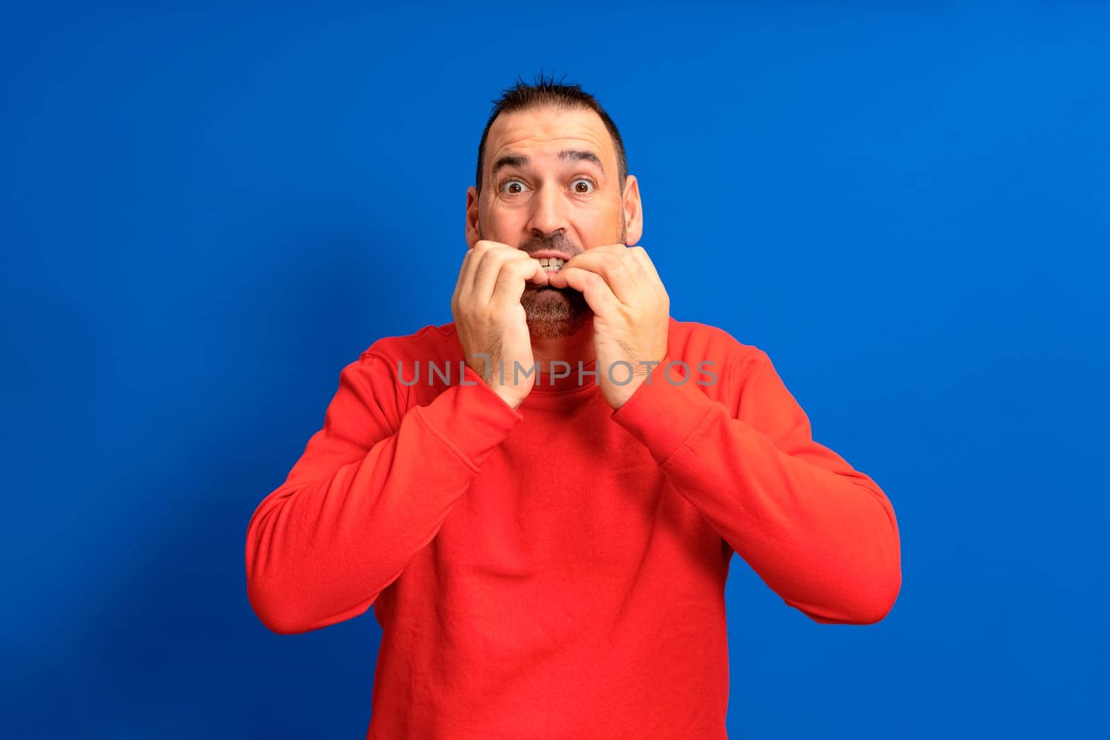 Handsome man wearing red jumper over isolated blue background looking stressed and nervous with hands on mouth biting nails. Anxiety problem.