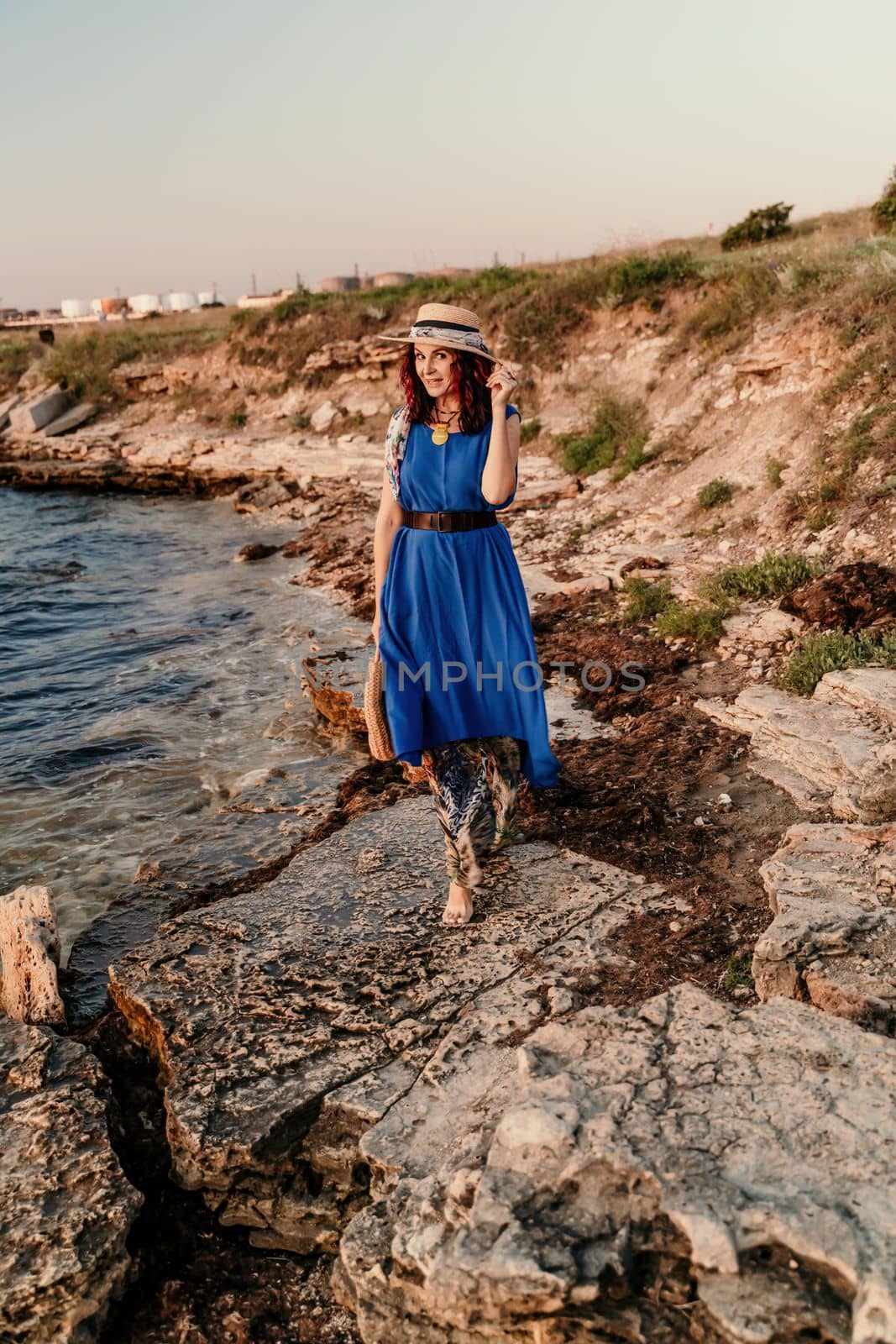 A woman in a dress, hat and with a straw bag is standing on the beach enjoying the sea. Happy summer holidays.