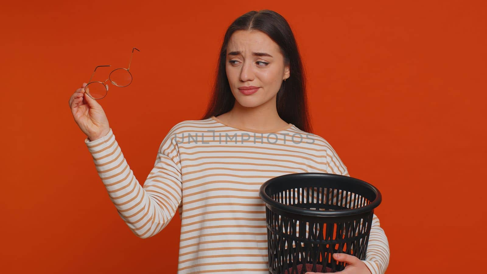 Lovely woman taking off, throwing out glasses into trash bin after medical vision laser treatment therapy surgery, looking smiling at camera. Young adult girl isolated alone on red studio background