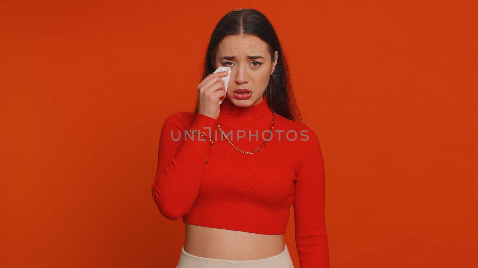 Upset disappointed pretty young woman in crop top wipes tears and cries from despair being sad because of unfair things. Millennial girl in depression, despair, loss isolated on red studio background