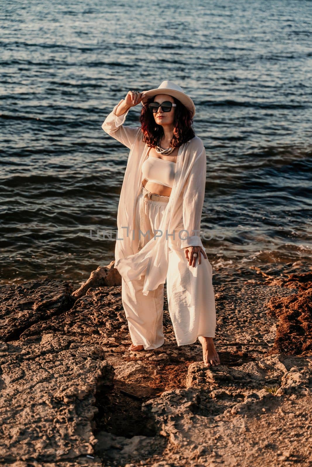 A woman in a white pantsuit and hat is standing on the beach enjoying the sea. Happy summer holidays.