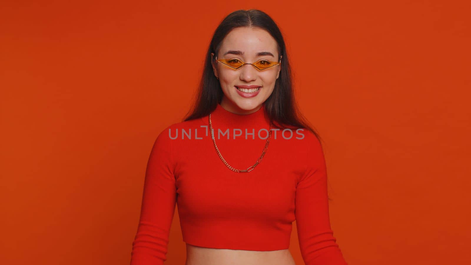 Portrait of happy pretty young woman in crop top wearing sunglasses, looking at camera with toothy charming smile, flirting, expressing optimism. Millennial girl isolated on red studio background