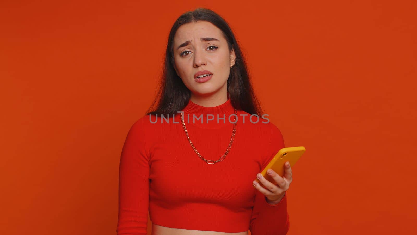 Pretty woman playing on smartphone typing browsing, loses becoming surprised sudden lottery results, bad fortune, loss, game fail, bankruptcy. Young girl isolated alone on red studio wall background