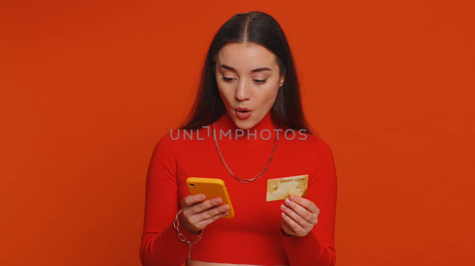 Sincere pretty woman customer using credit bank card and smartphone while transferring money, purchases online shopping, payment. Finance and internet. Young girl isolated alone on red background
