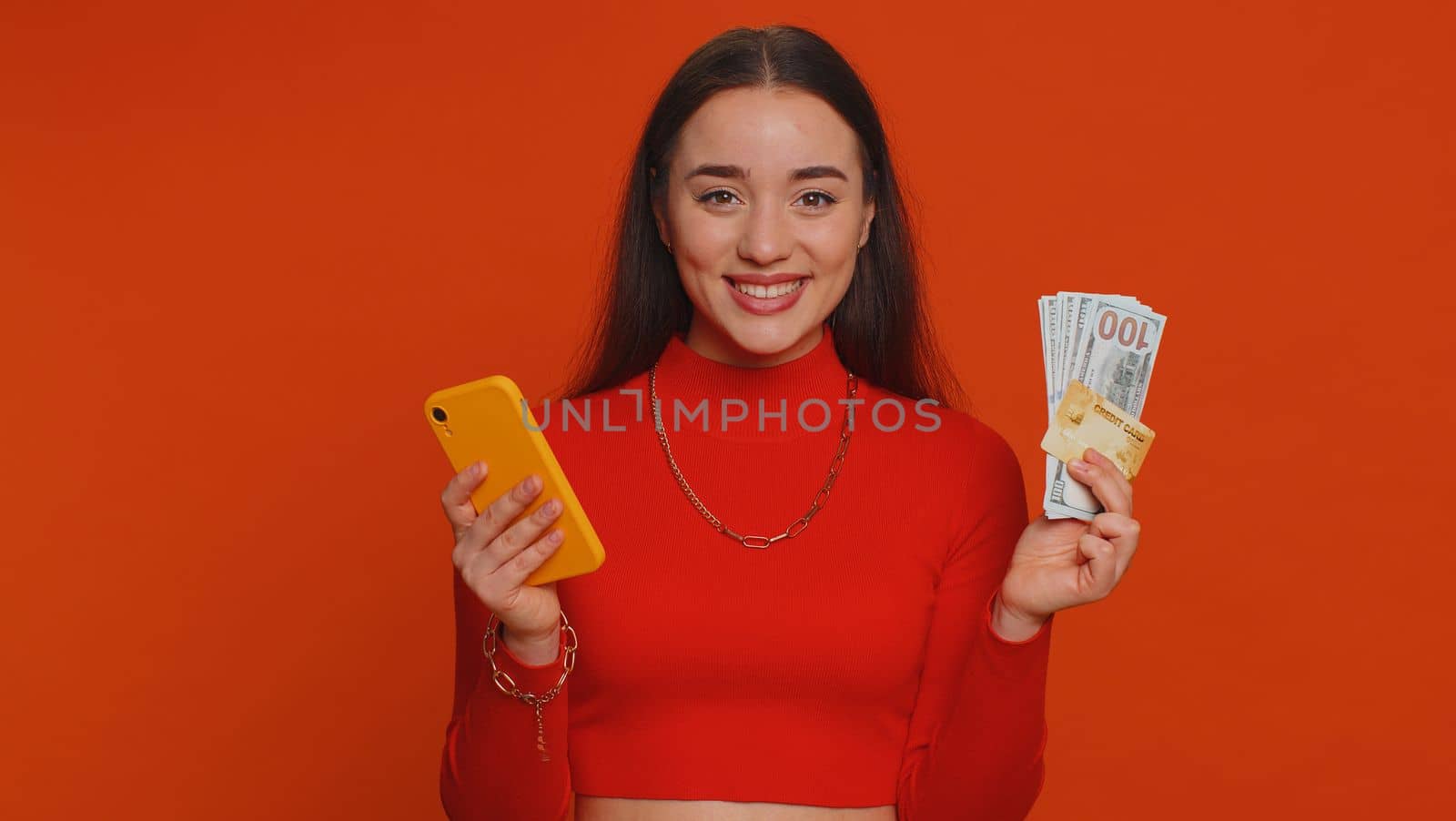 Sincere one woman customer using credit bank card and smartphone while transferring cash money, purchases online shopping, payment. Finance and internet. Young girl isolated alone on red background