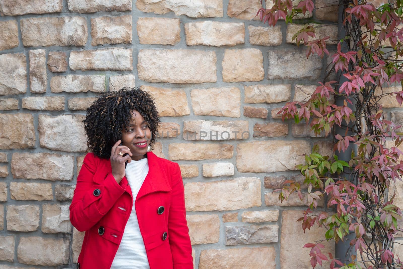 African american woman in red coat talking on smartphone near brick wall by KaterinaDalemans