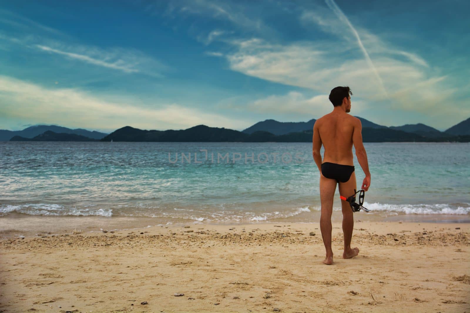 Full body shot from the back of a handsome young man standing on a beach in Phuket Island, Thailand, shirtless wearing boxer shorts, showing muscular fit body