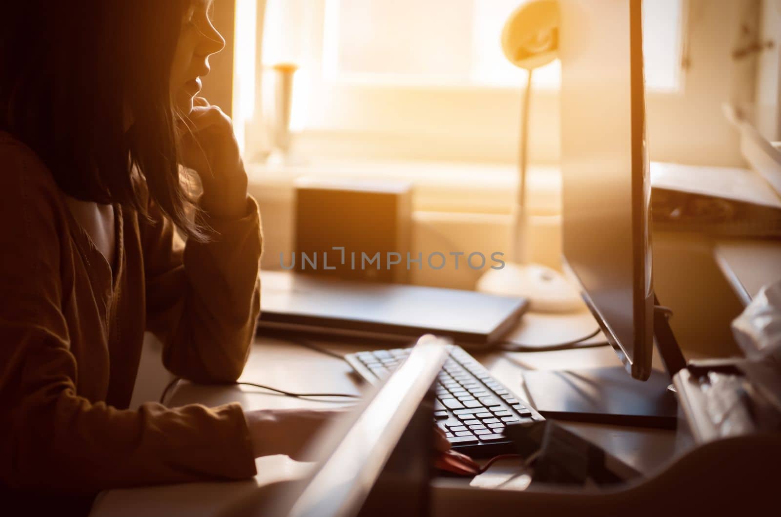 A young girl sits at home by the window and works online, studies, develops a business and her creative project, a woman types on the keyboard and looks at the monitor in the rays of the sun.