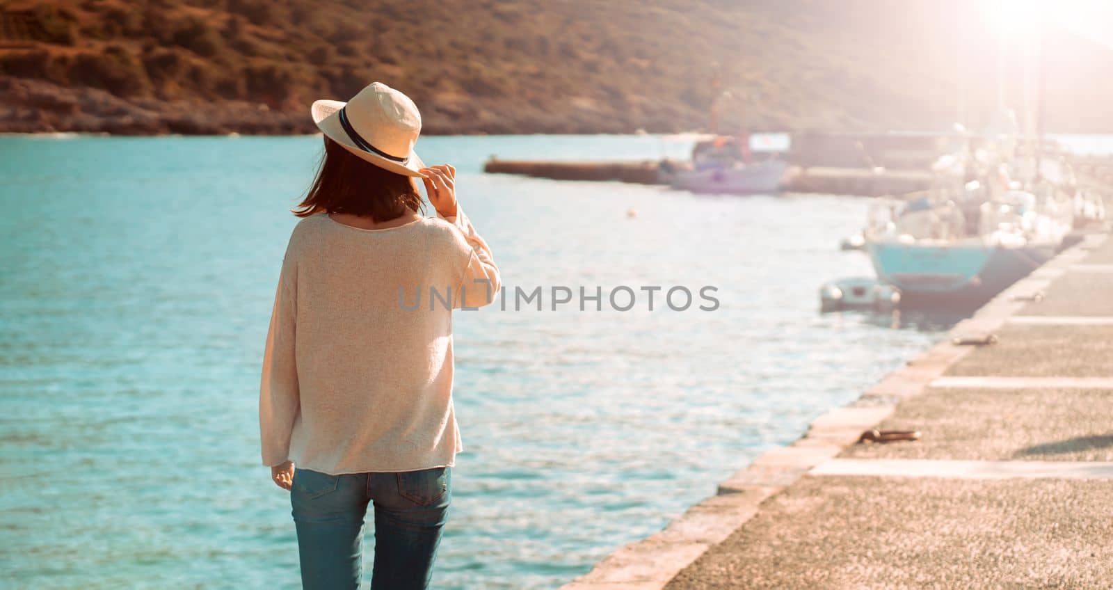 A young girl in a hat looks at the picturesque view with the sea and moored yachts on a sunny day, a backpacker travels and discoveries beautiful places on the coast during her holidays.