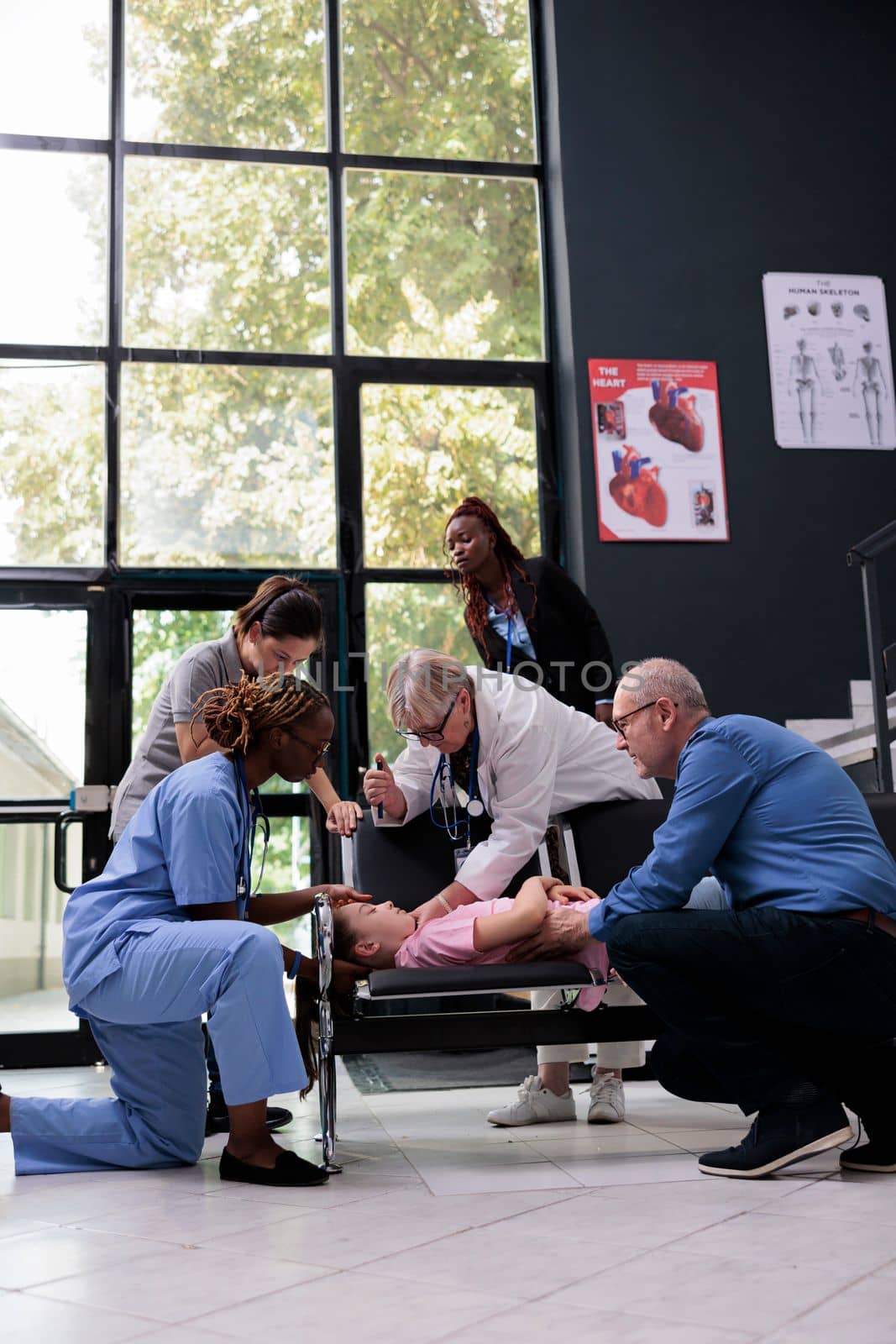 Scared family after unconscious little child falling down on chair in hospital waiting room by DCStudio