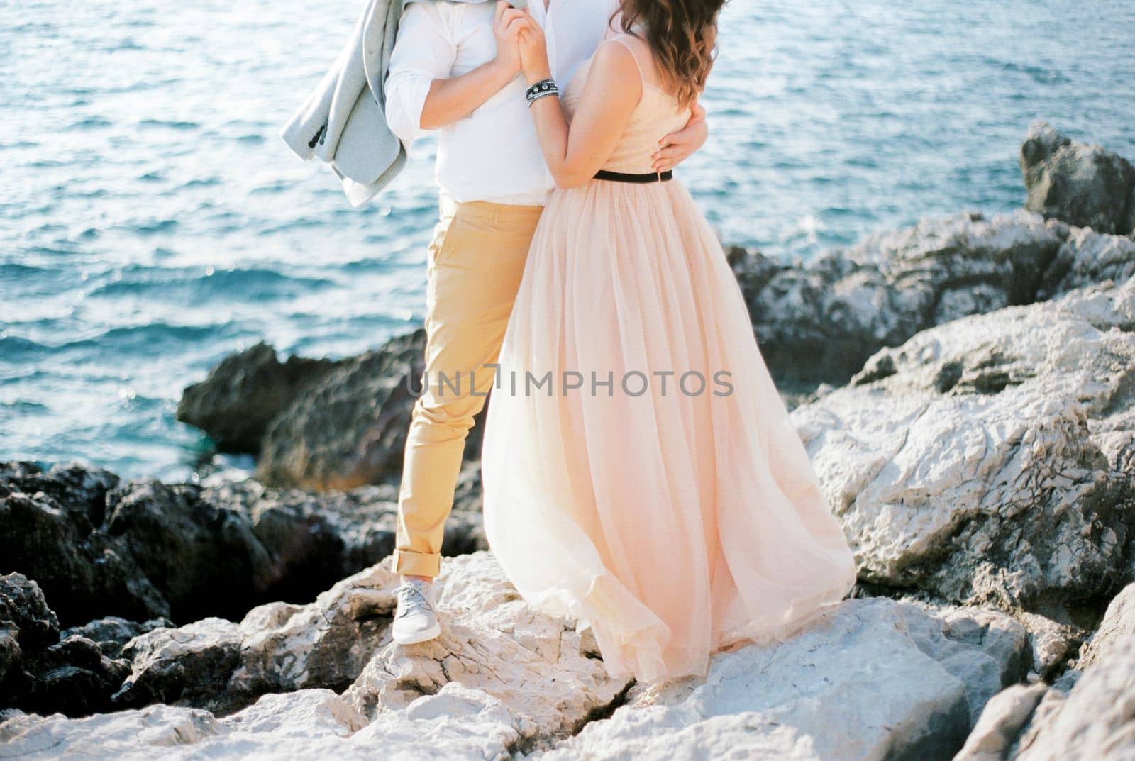 Groom with a jacket on his shoulder hugs bride on a rocky seashore. Cropped. High quality photo
